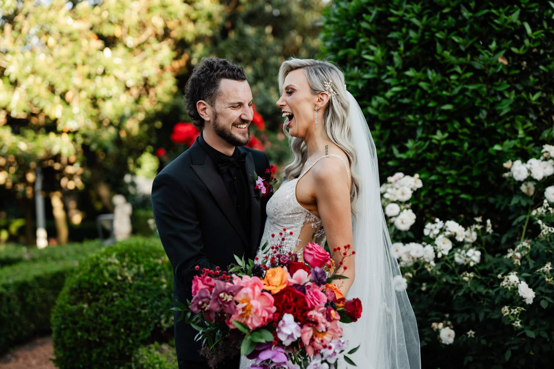 groom wearing black suit and bride in white dress holding pink red purple flowers laughing at wedding in Perth