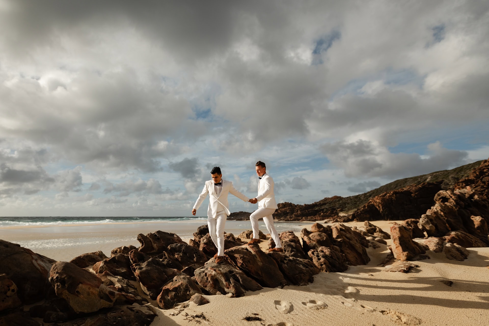 two grooms in white suits holding hands walking on rocks