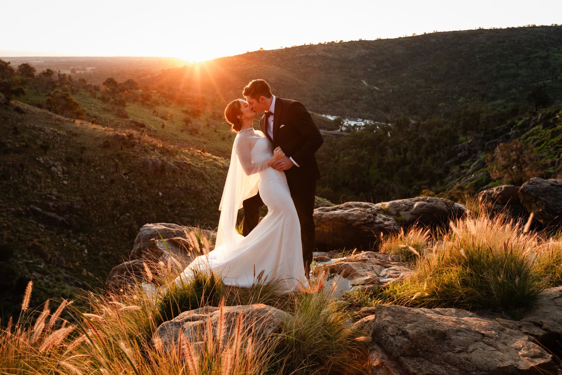 Perth wedding photographer bride in white dress and groom in black suit with bow tie kissing on cliff at sunset in Perth