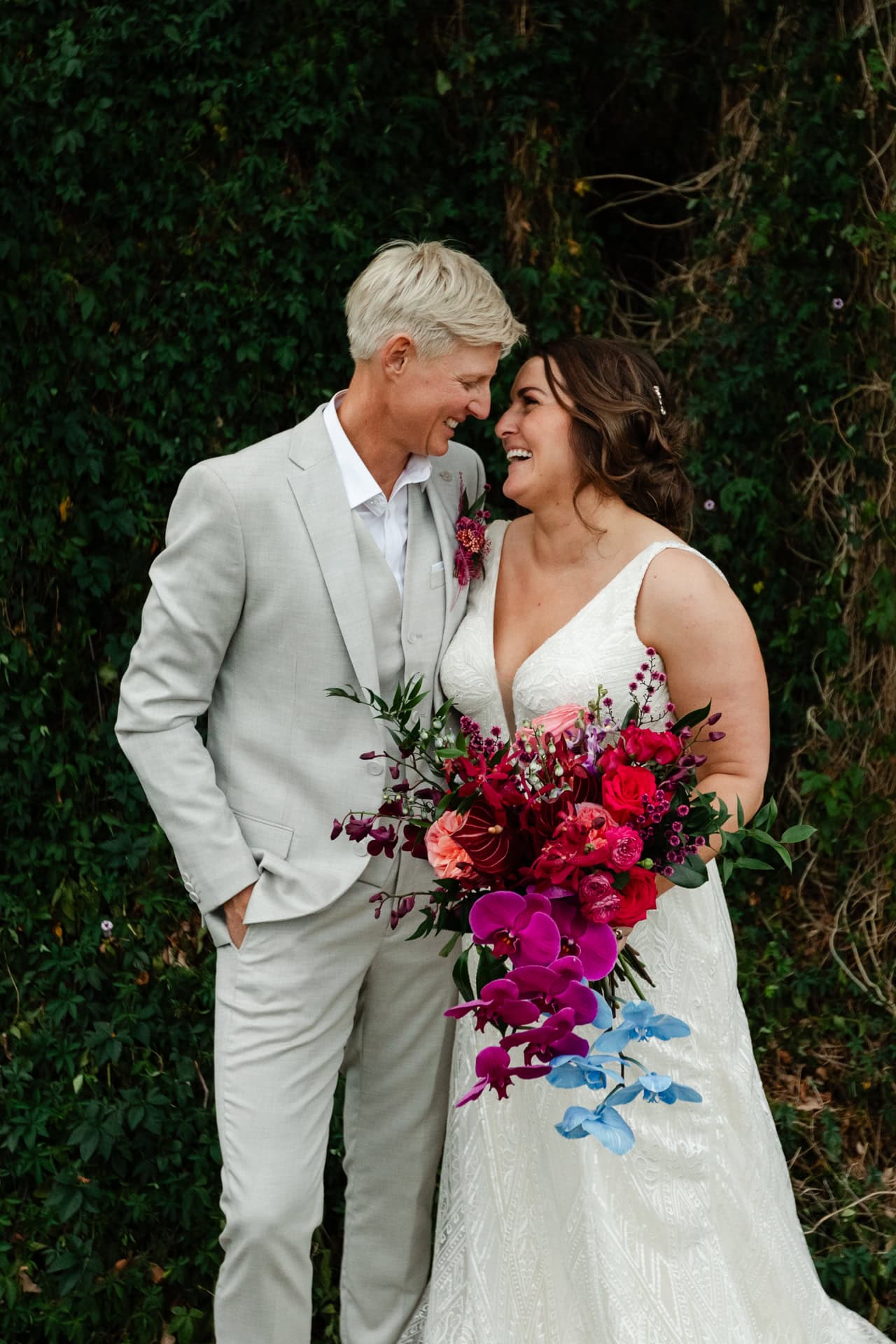 two brides one in cream suit hugging other bride in white dress laughing