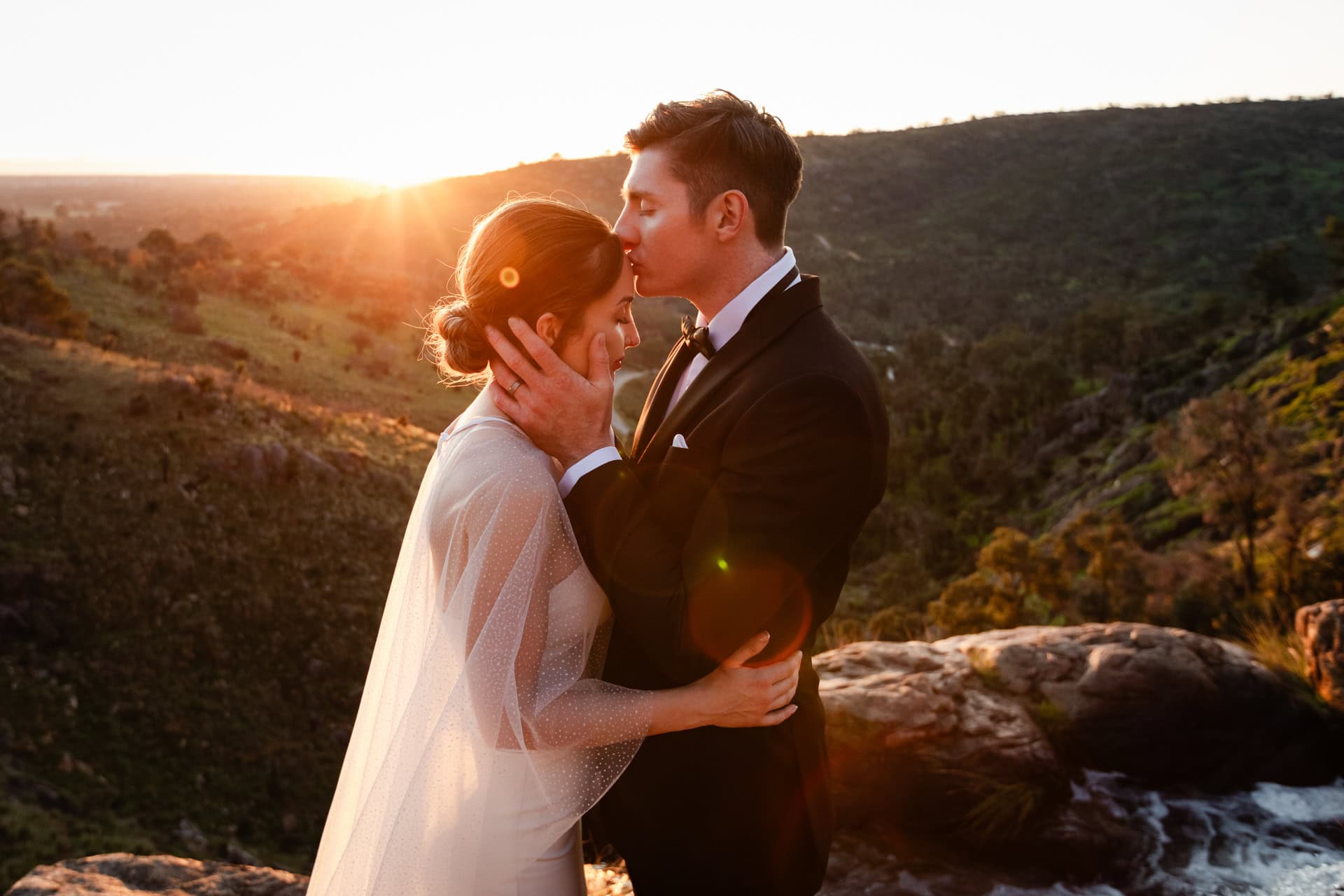 groom kissing bride on head at sunset on cliff