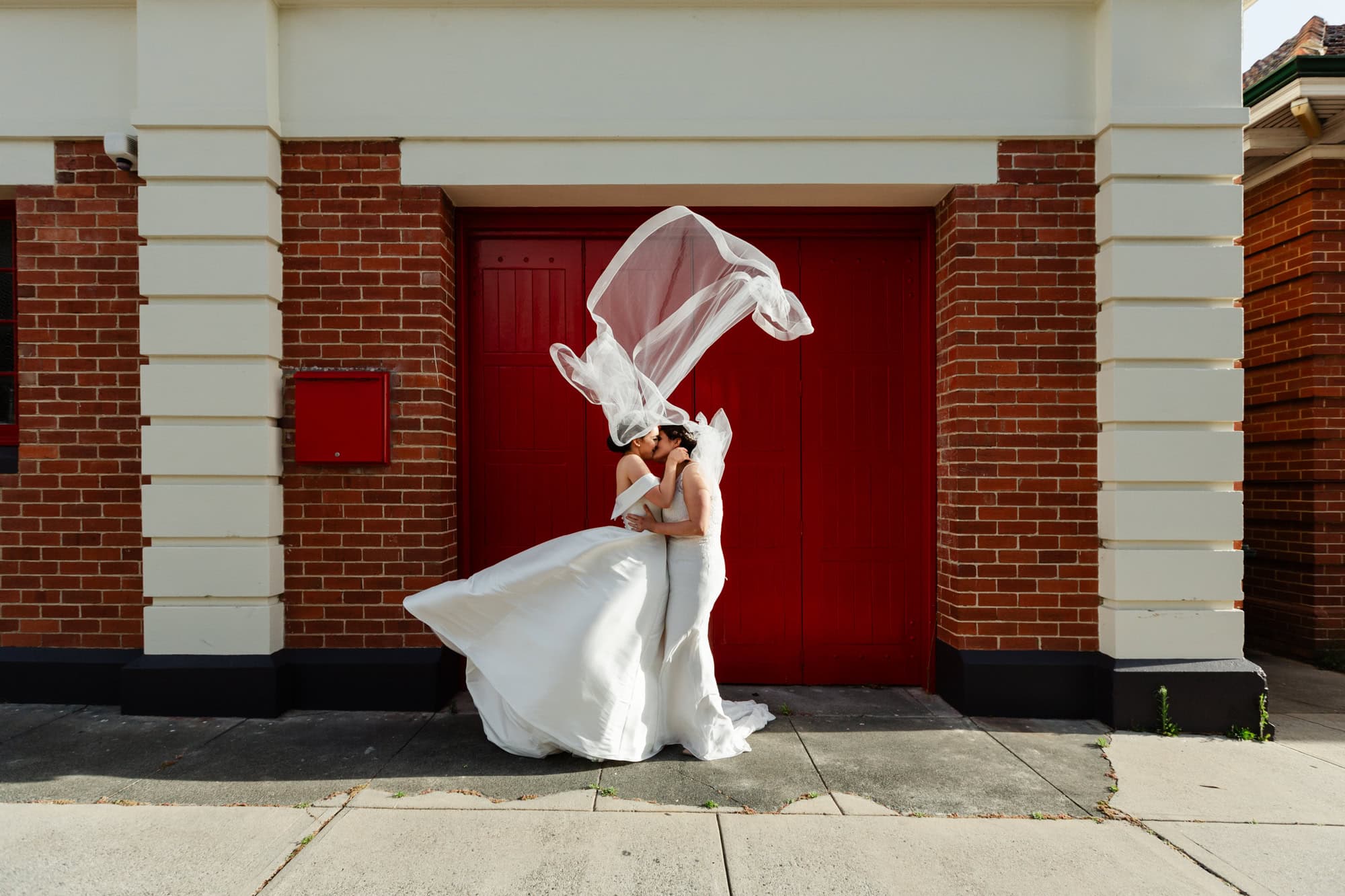 wedding photos Perth two brides wearing white dresses kissing in front of red door with veil flying above in air