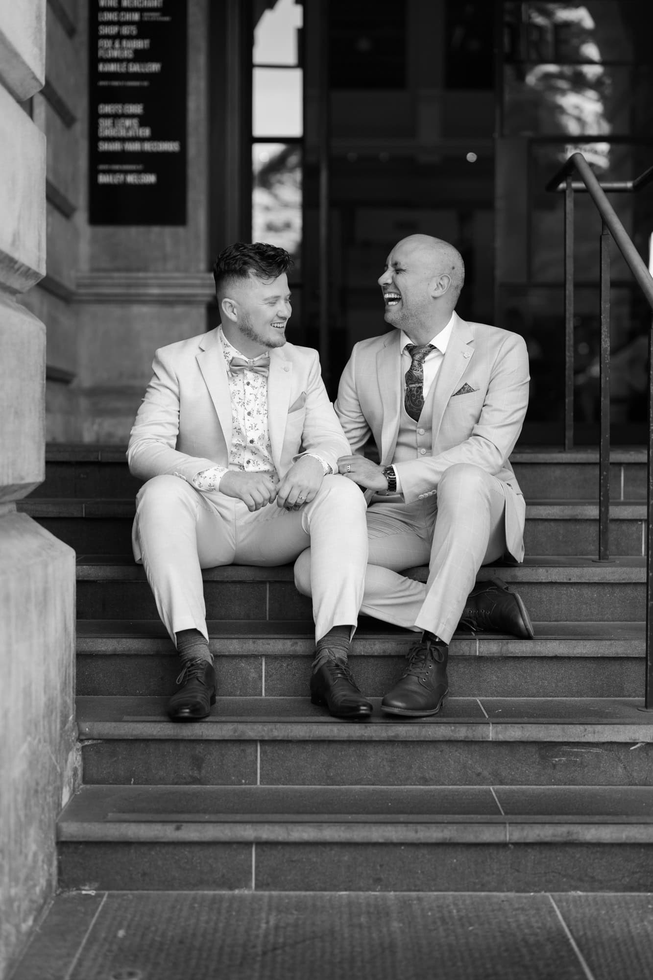 wedding photos Perth two grooms wearing suits sitting on steps laughing