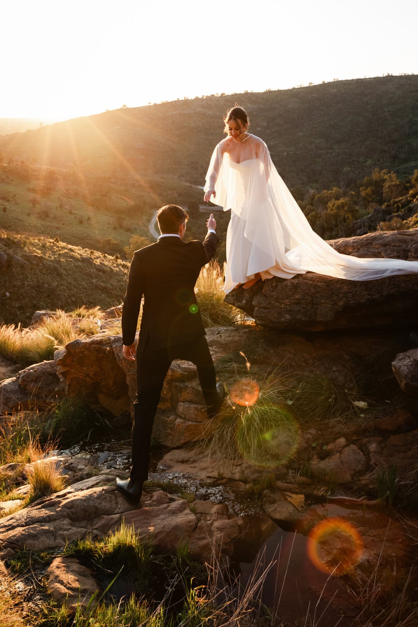 wedding photos Perth groom wearing black suit reaching for hand of bride wearing white dress and no shoes standing on rock at sunset