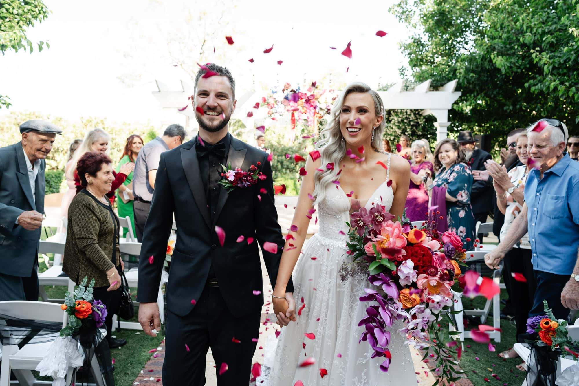 wedding photos Perth groom wearing black suit and bride wearing white dress walking down ceremony aisle with red pink petals flying through air