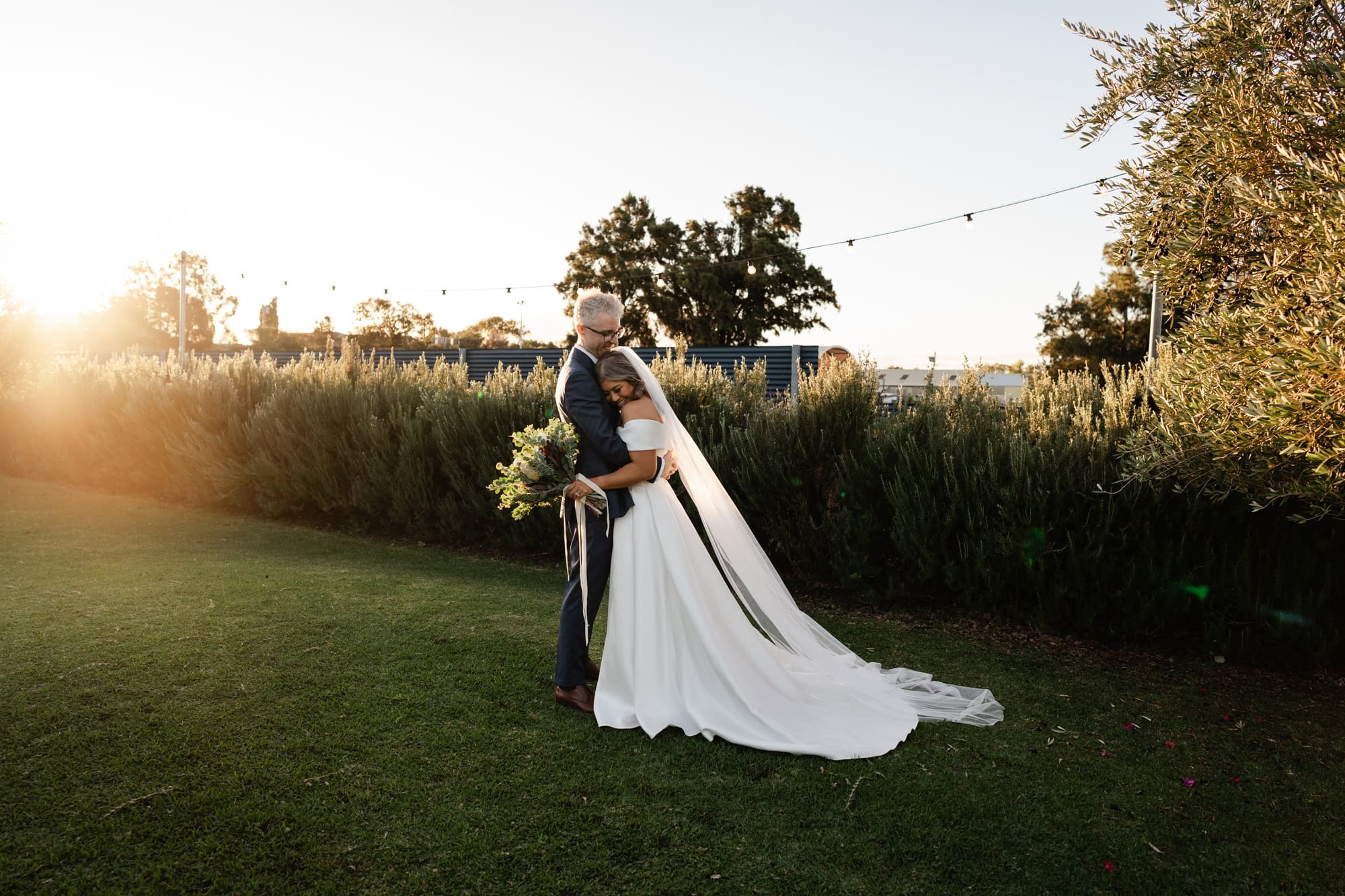 wedding photos Perth groom wearing blue suit hugging bride wearing white dress on grass hill at sunset