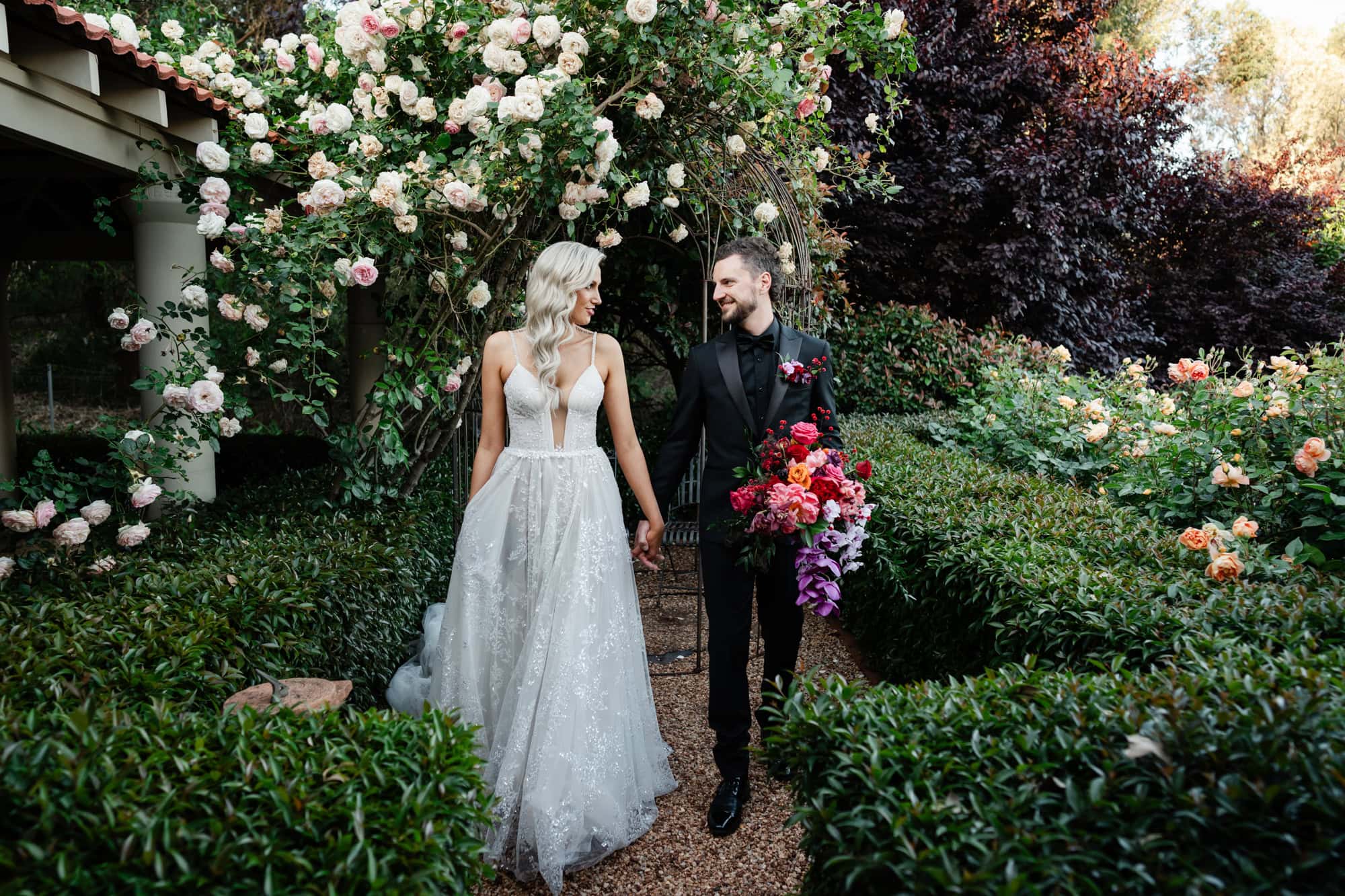 wedding photos Perth groom wearing black suit carrying red purple pink bouquet and holding hand of bride wearing white dress in garden