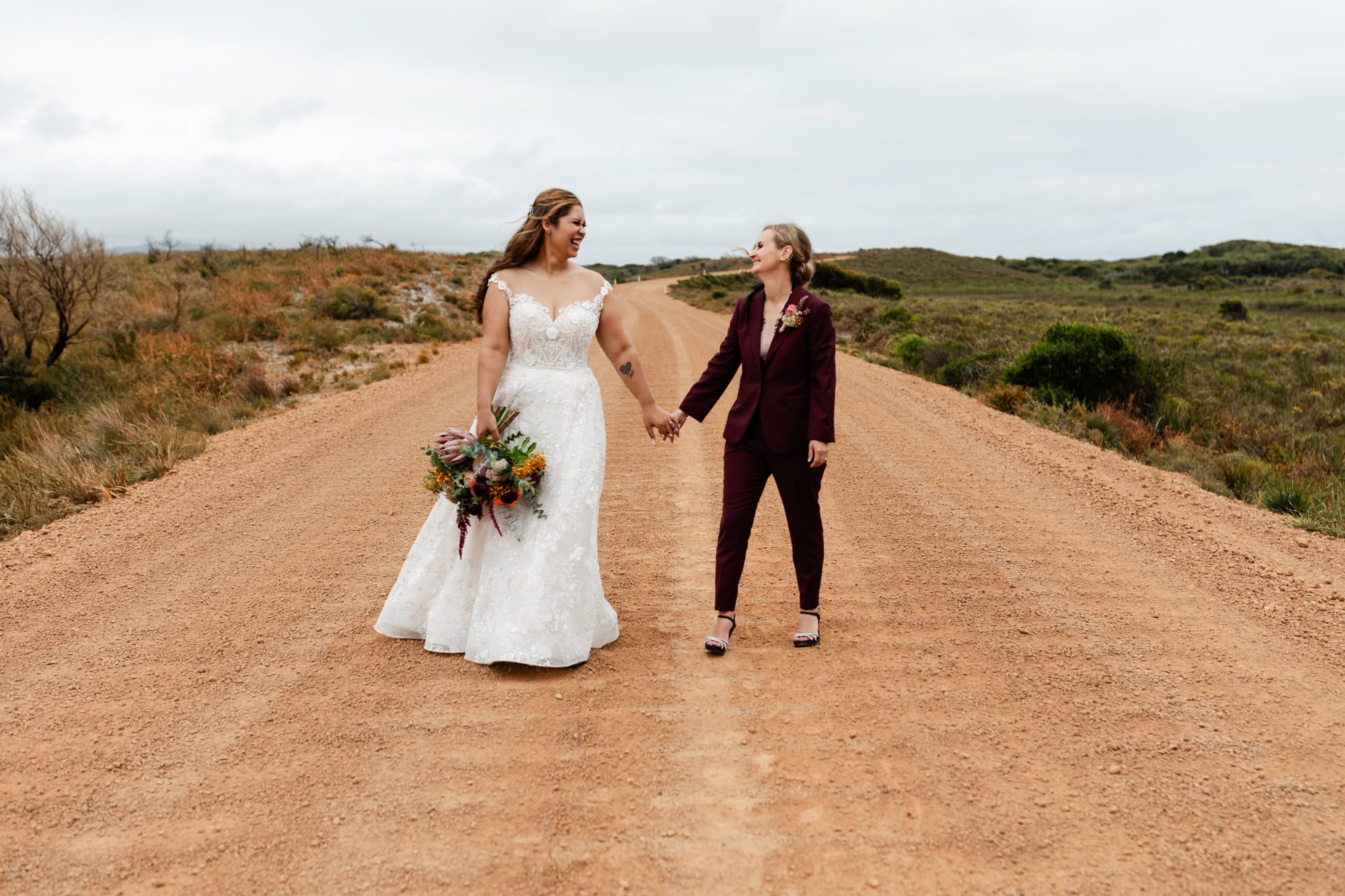 wedding photos Perth bride wearing white dress holding maroon bouquet and holding hands with bride in maroon suit standing on dirt orange road laughing