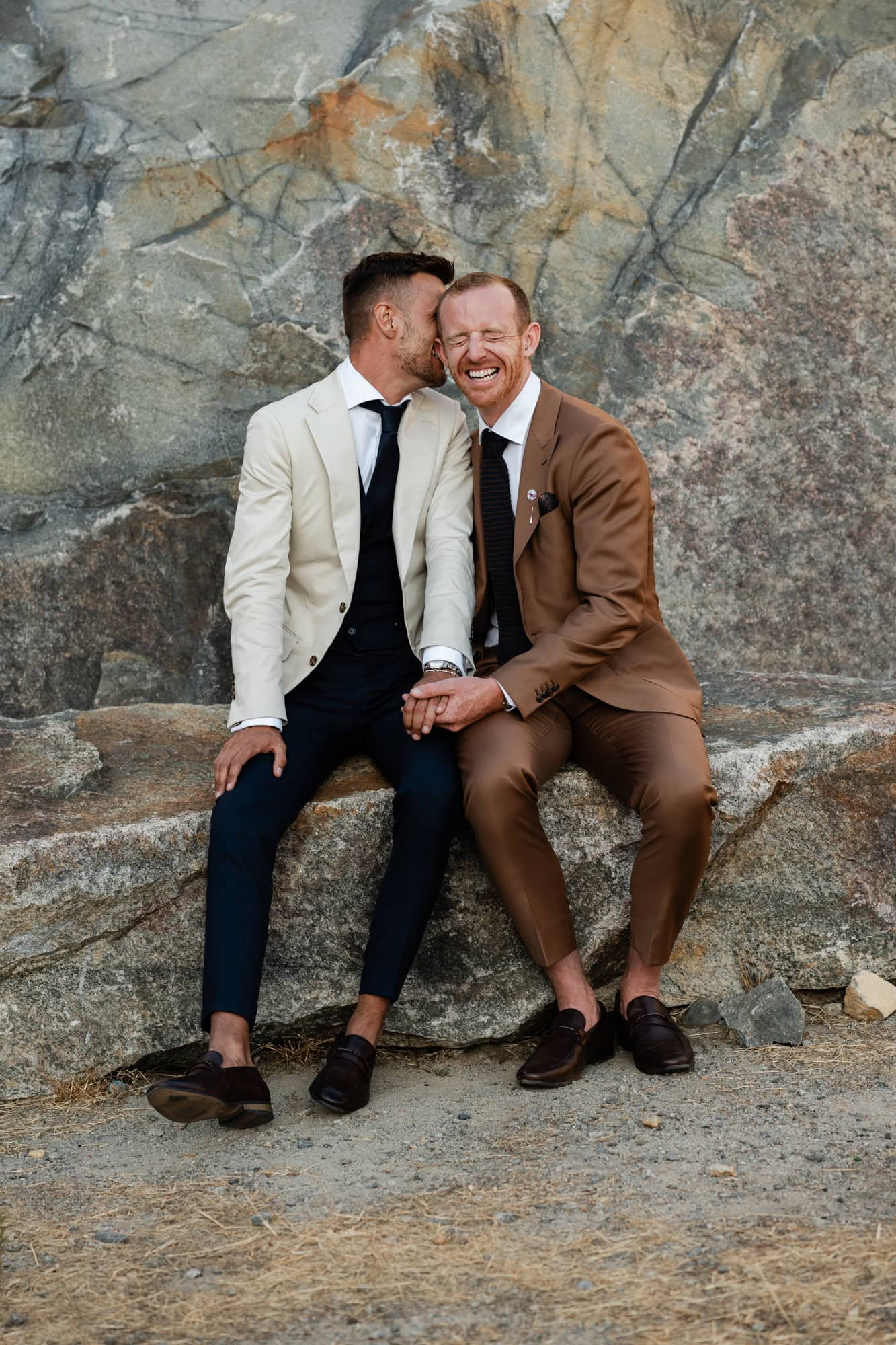 wedding photos Perth groom in suit with cream jacket sitting on rock next to groom in brown suit hugging laughing